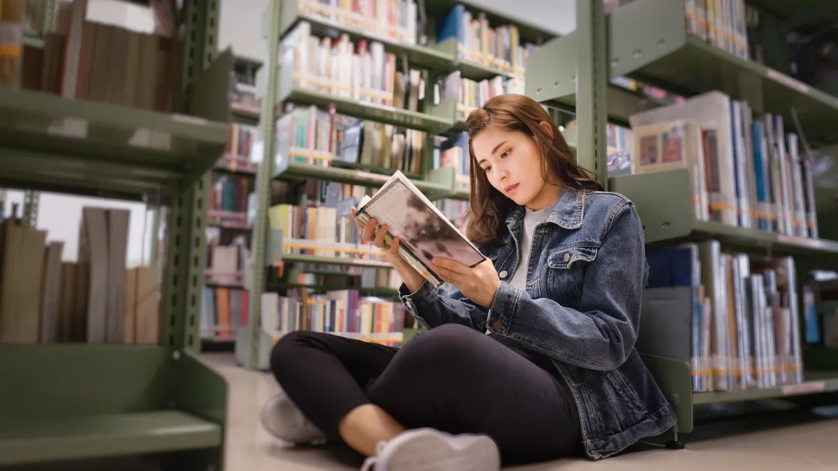 Student sits on floor in a library holding a book. Behind the student are rows of bookshelves.