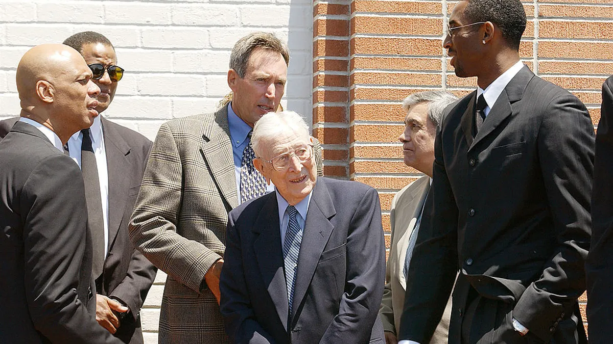 Former NBA player Kareem Abdul-Jubbar, former UCLA coach John Wooden, and NBA player Kobe Bryant stand in a group outside a brick building.