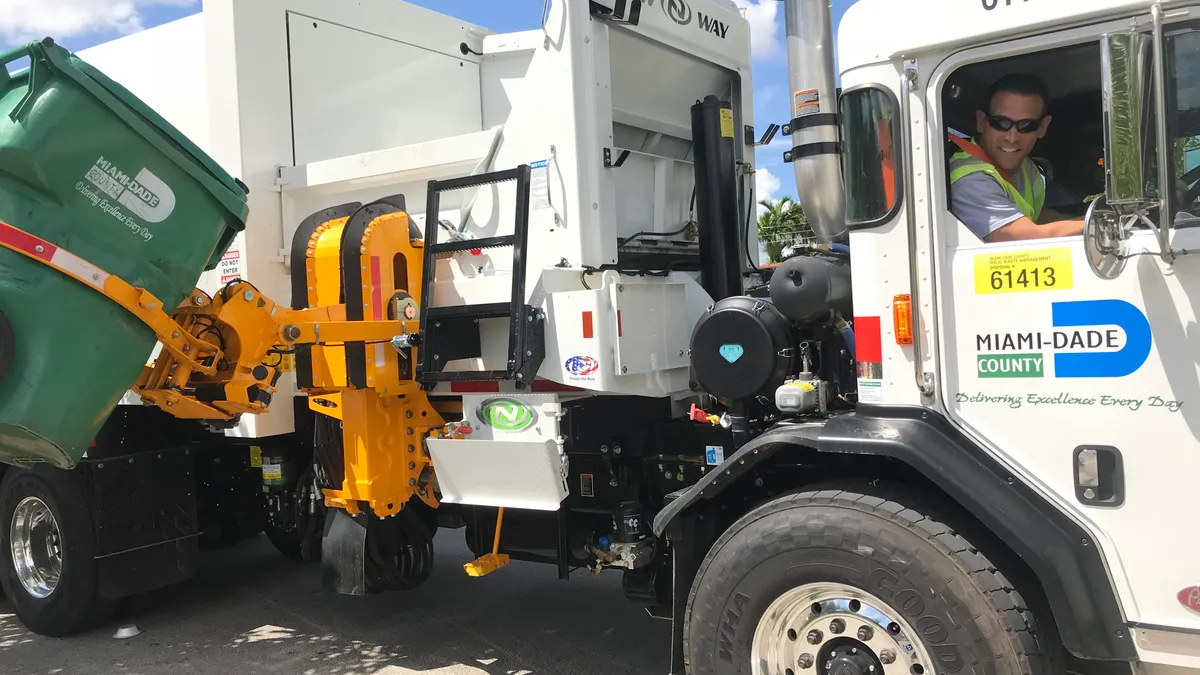 A man operates a side-loading refuse collection truck lifting a curbside waste bin. The truck's driver door is emblazoned with the Miami-Dade County Department of Solid Waste Management logo.