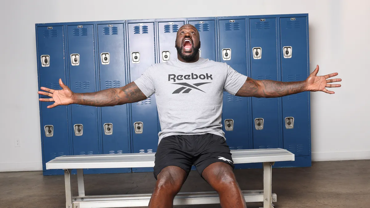 Shaquille O'Neal sitting on a bench in a Reebok shirt with his arms flung wide.