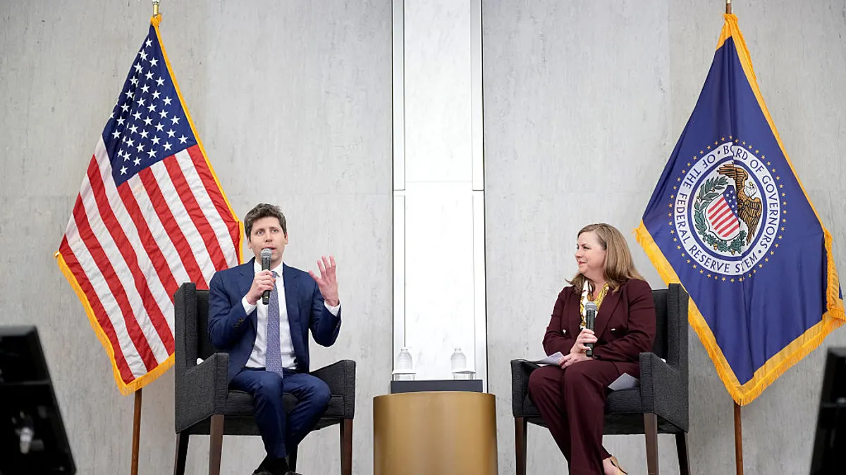 Sam Altman sits on the left side of a stage, in front of an American flag, while speaking into a microphone and gesturing, as Michelle Bowman sits on the right side of the stage in front of a Federal Reserve flag.