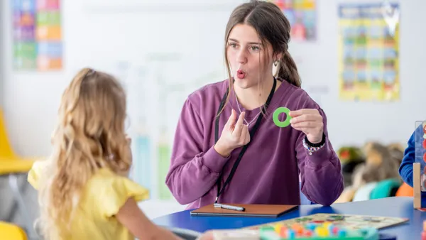 An adult is in a classroom and is sitting at a table holding the letter "O" and curving their mouth into an O. A young student is in front of the adult with their back to the camera.