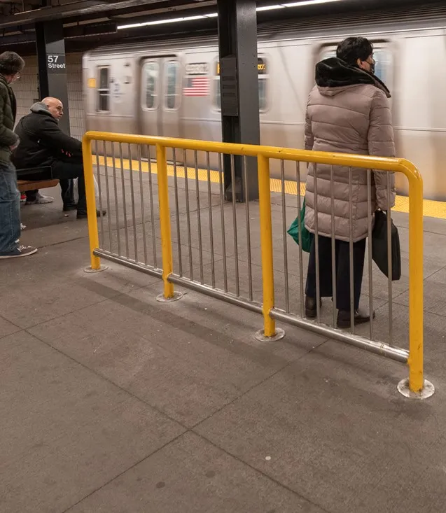 A short yellow metal fence is mounted to a subway platform in New York City as a silver train pulls into the station with two standing and one seated person.