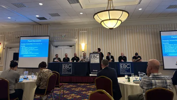 A group of seven people sit at tables on a stage, with one standing at a lectern in the middle, at an Environmental Business Council event.