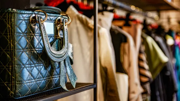 In the foreground, there is a black quilted handbag. In the background is a blurred rack of clothing. It appears to be a clothing store.