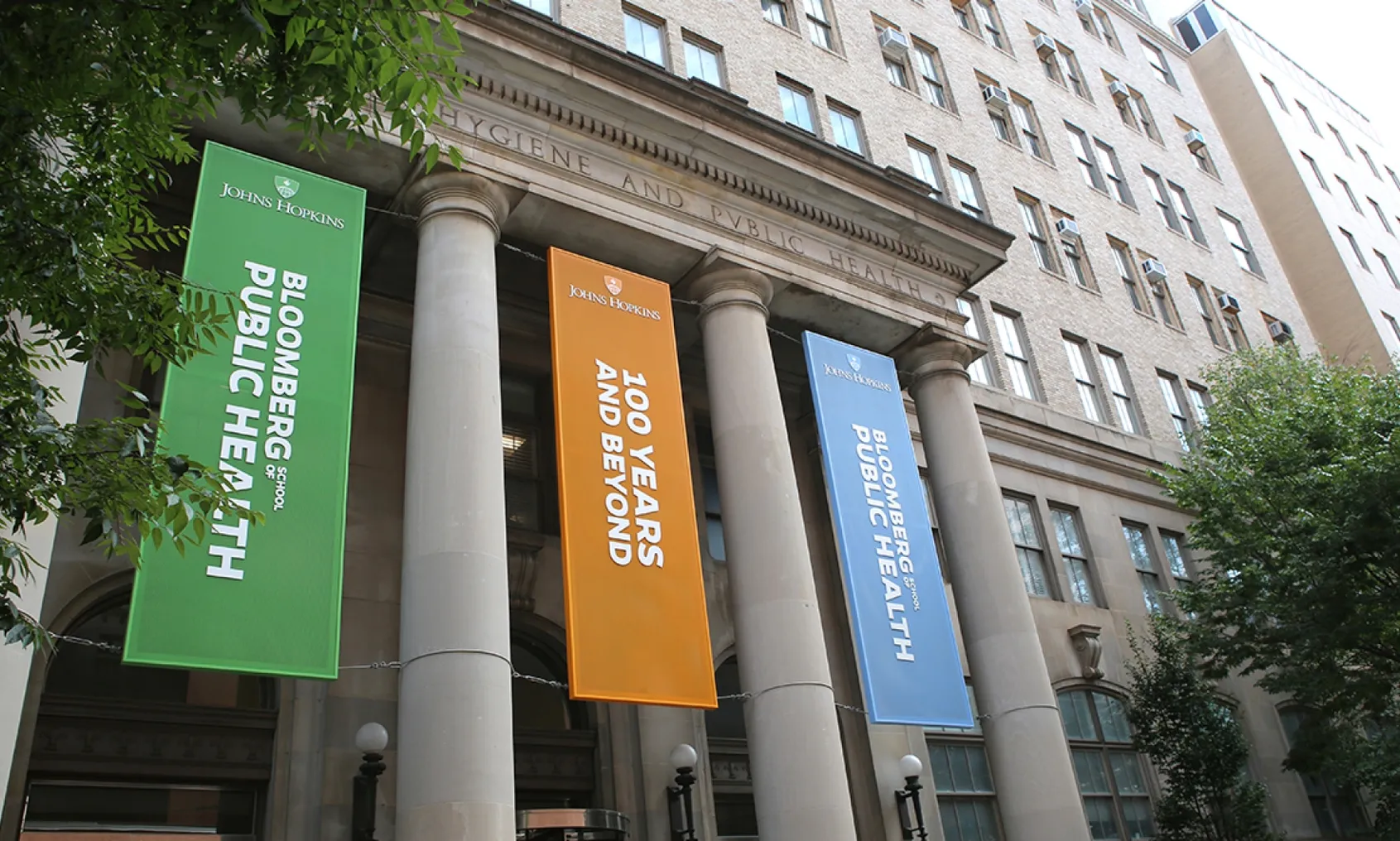 The front of an academic building with coliseum pillars. Between the pillars are hanging banners in orange, green and blue.