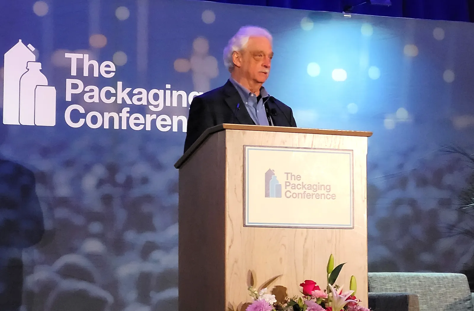 A person stands at a podium in front of a blue background that has a logo for The Packaging Conference.