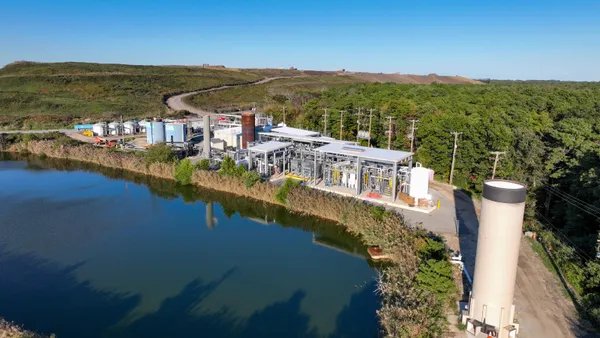 Aerial view of an industrial facility with tanks and a complex system of pipes. A small pond is in the foreground, and a road leads to a landfill in the background.