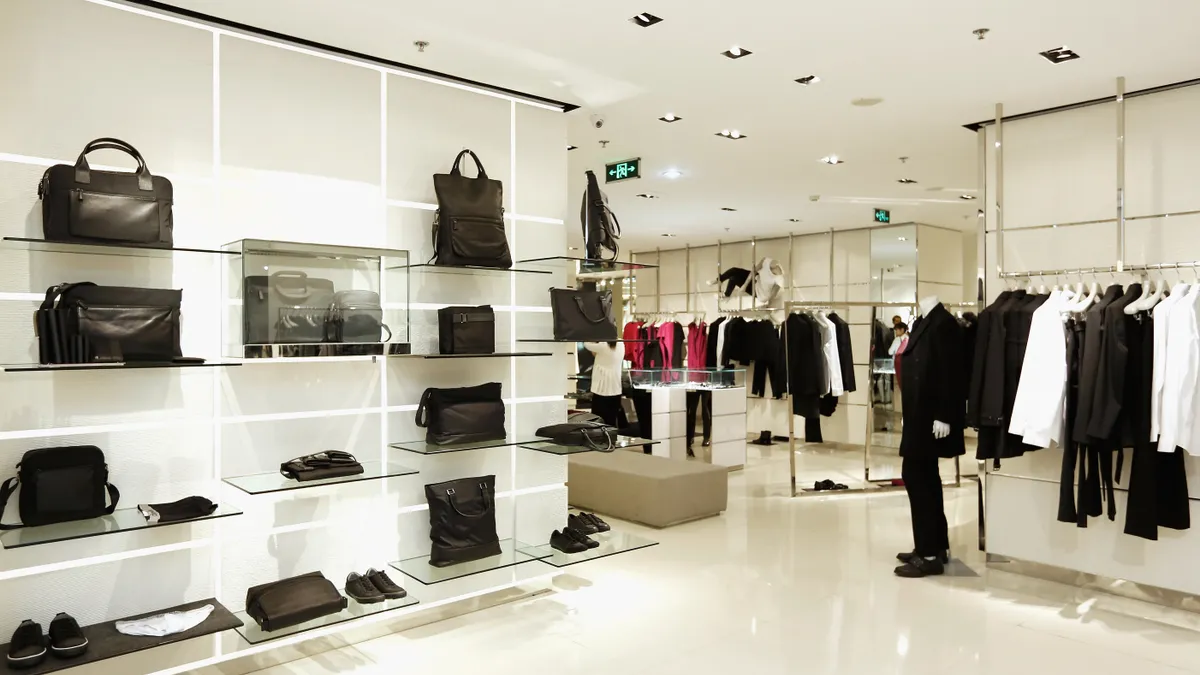 The interior of a Calvin Klein store. There are shelves displaying handbags and shoes as well as racks and mannequins featuring suiting.