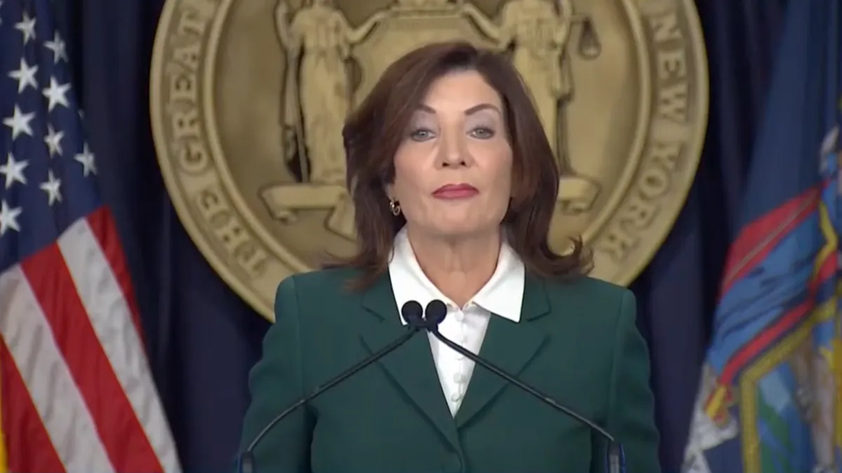 Woman in green suit at a podium with microphones with the seal of the state of New York behind her.