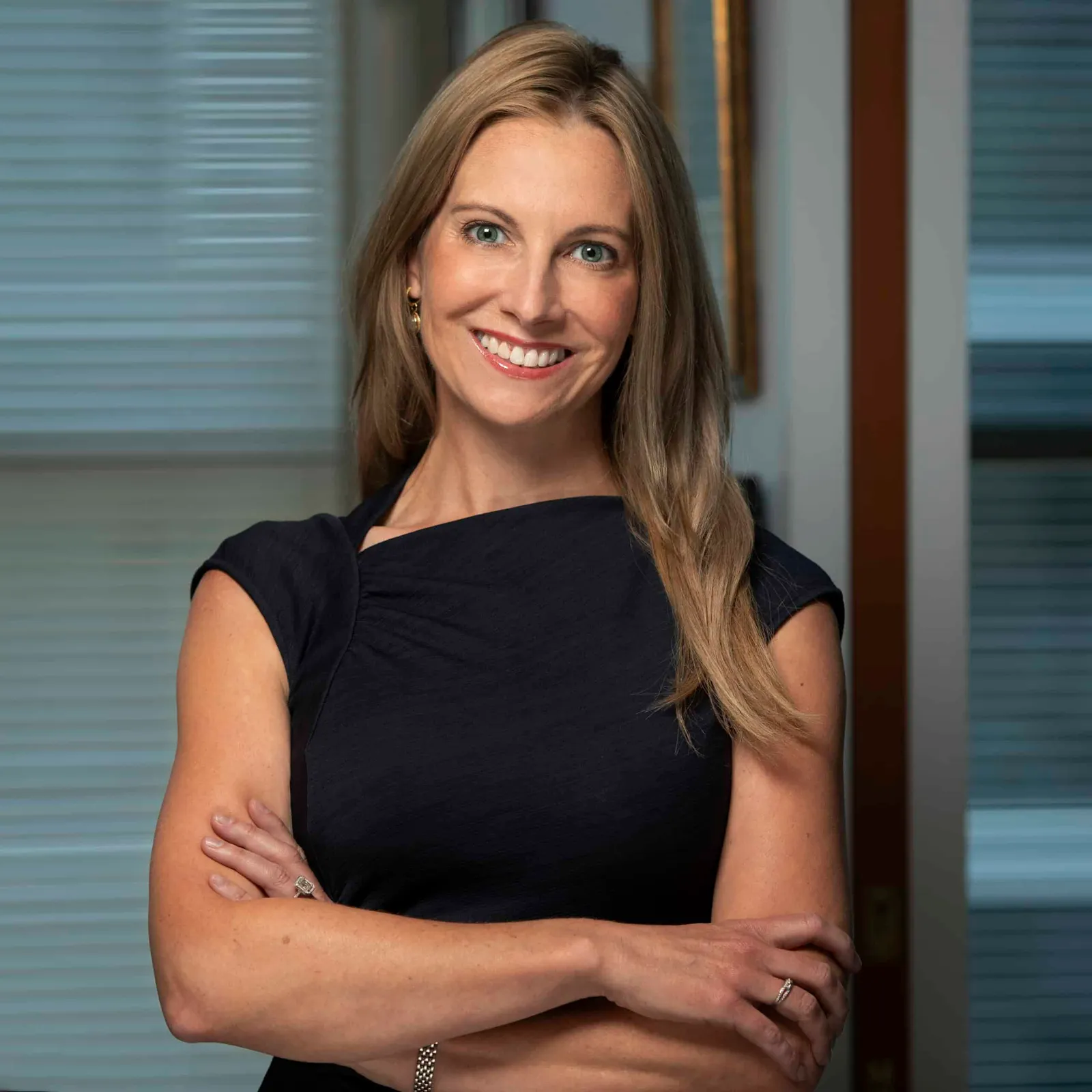 A blonde woman wearing a black dress with arms folded over her torso.