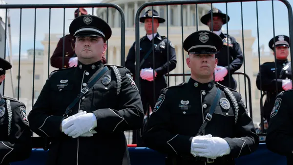 Officers wearing black uniforms and white gloves stand with hands folded in front of them and the U.S. Capitol in the background.