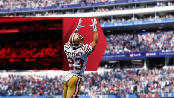 A football player in a white, gold and red uniform is shown in the air with arms up, ready to catch a football. A stadium crowd is blurred in the background, and a red shield logo is seen in the middle of the image.