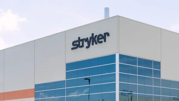 A modern industrial building with beige and white walls and large blue-tinted glass windows. The name “Stryker” is mounted in black letters on the upper portion of the building. A white smokestack protrudes from the roof, and the sky is light and partly cloudy.