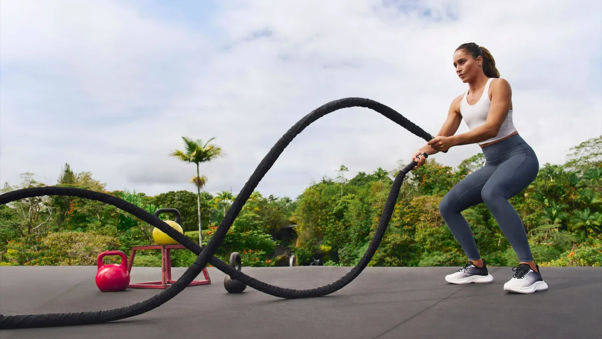 A person with a ponytail holds two thick exercise ropes and wears a cropped workout top and leggings.