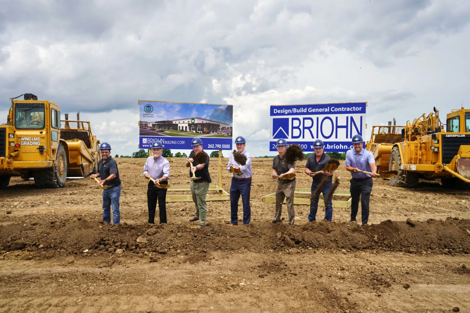 Seven people wearing hard hats and holding shovels participate in a groundbreaking ceremony in front of construction equipment.