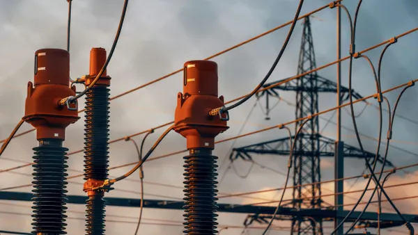 Electrical substation components and power lines with a transmission tower in the background at sunset.