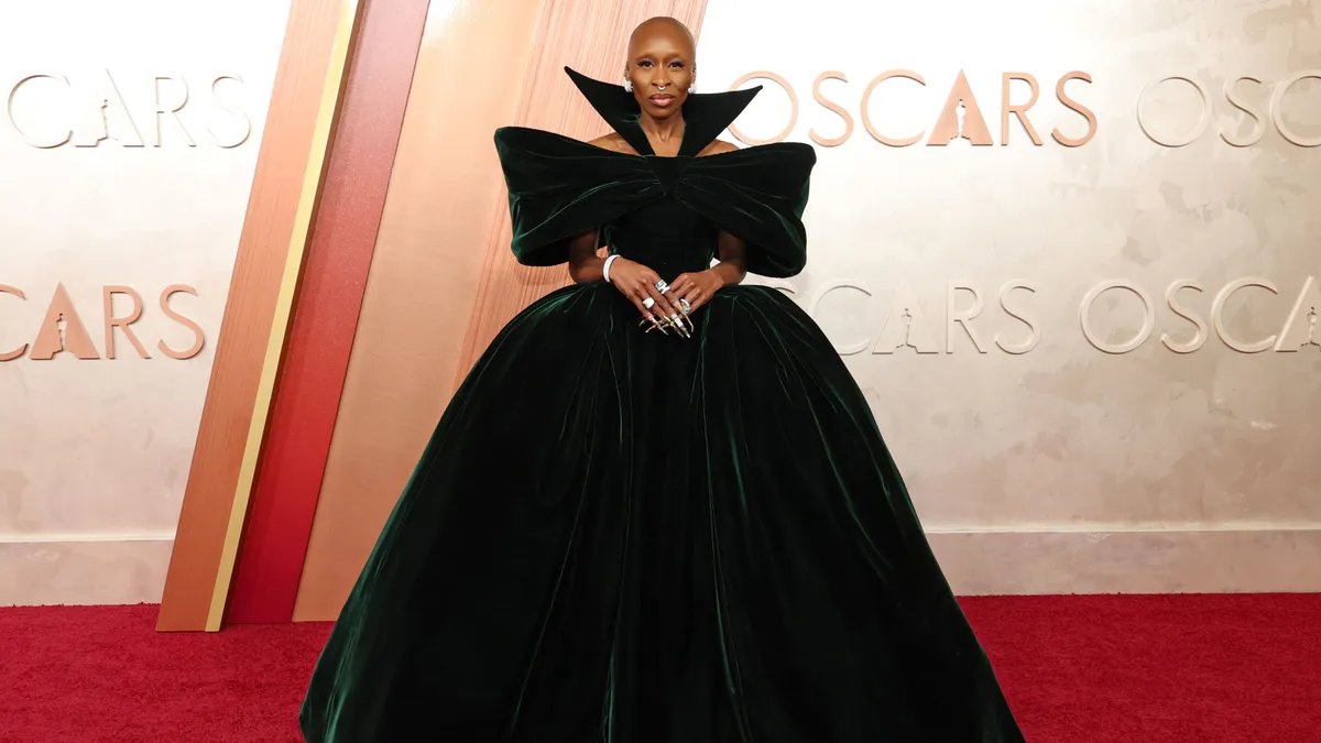 A woman in a green velvet dress with exaggerated shoulders and a wide skirt stands on a red carpet.