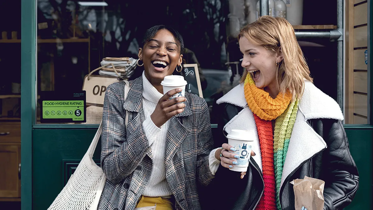 Two women in colorful winter clothes laugh joyfully outside a café, holding coffee cups. A food hygiene rating sign is visible.