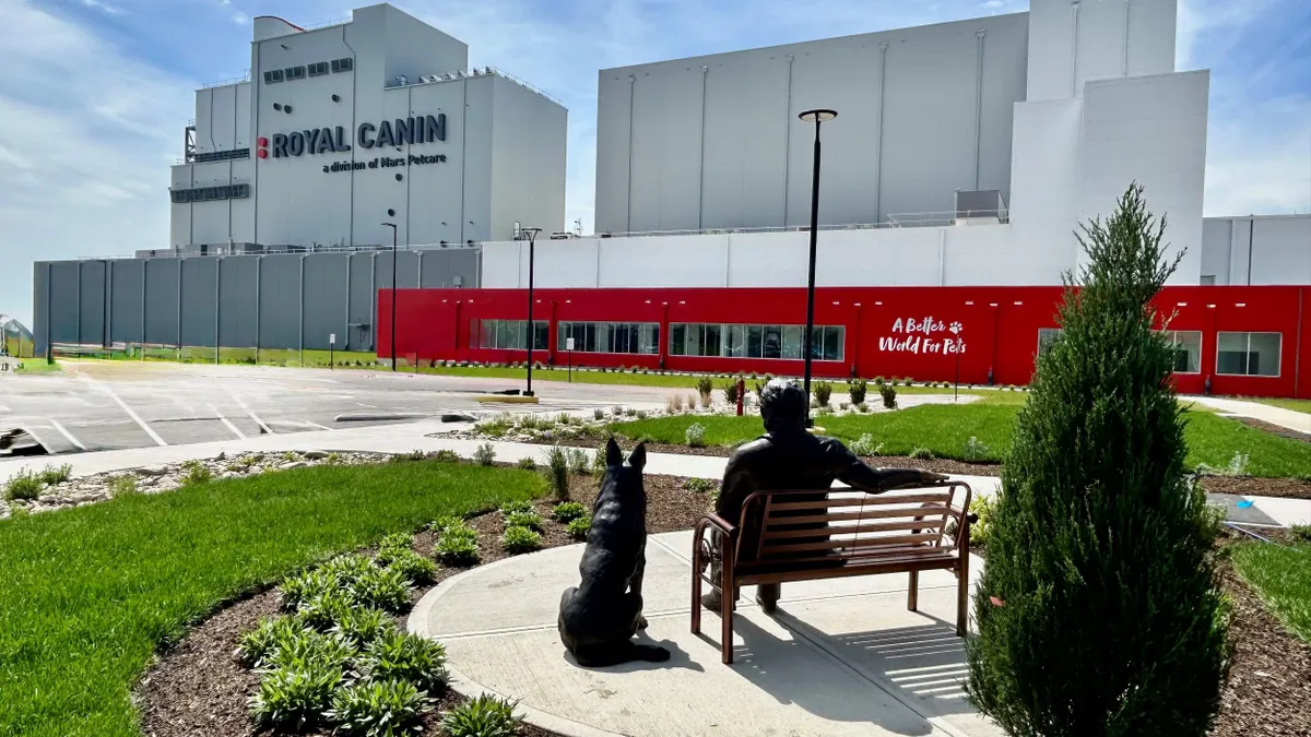 Statues of a person and a dog facing a gray and red factory.