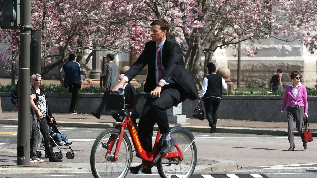 A man in a suit riding a bicycle along a street lined with cherry blossom trees.