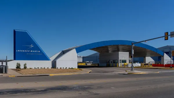 A blue and white Lockheed Martin sign on the left of a blue archway that's over a white parking attendant booth.