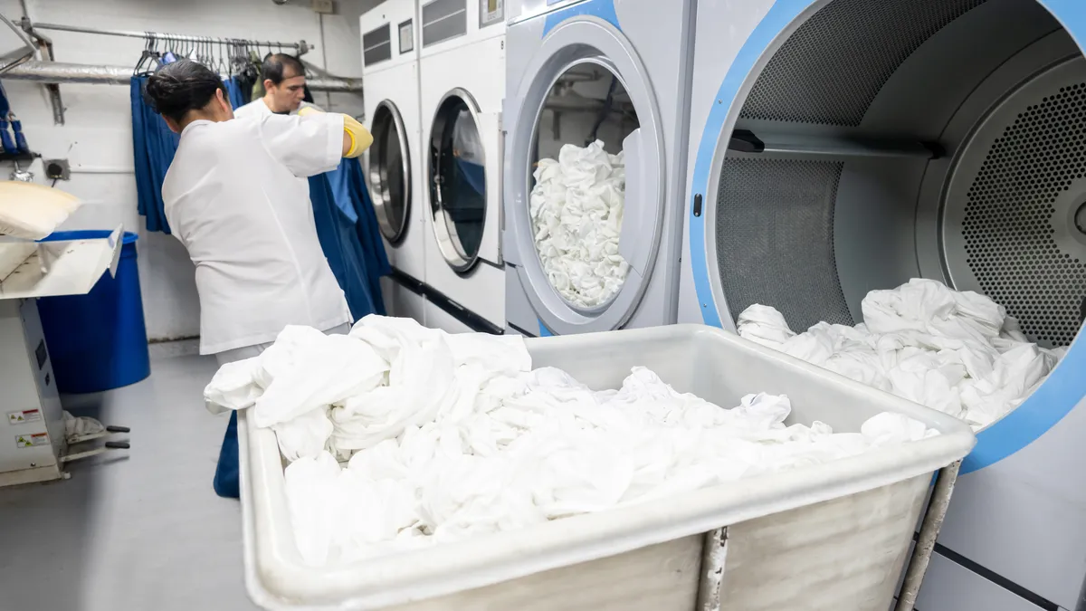 Two workers in white clothes load white towels into a front-load washing machine at a laundry facility.