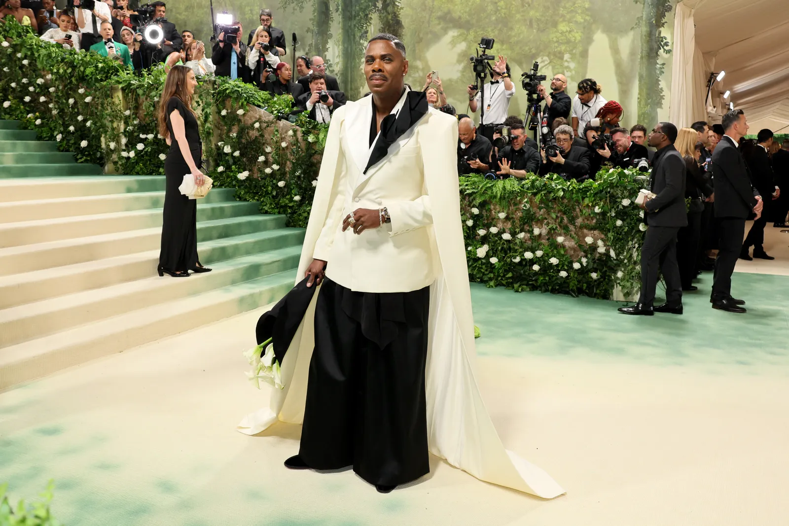 A person in a white and black suit holds calla lilies and stand in front of photographers.