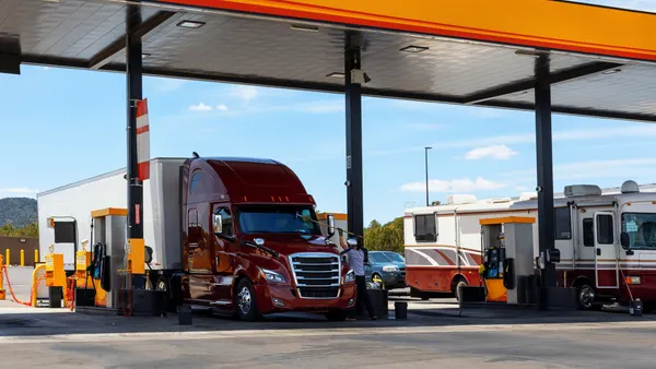 A truck driver washes the windshield of a tractor cab next to fuel pumps.