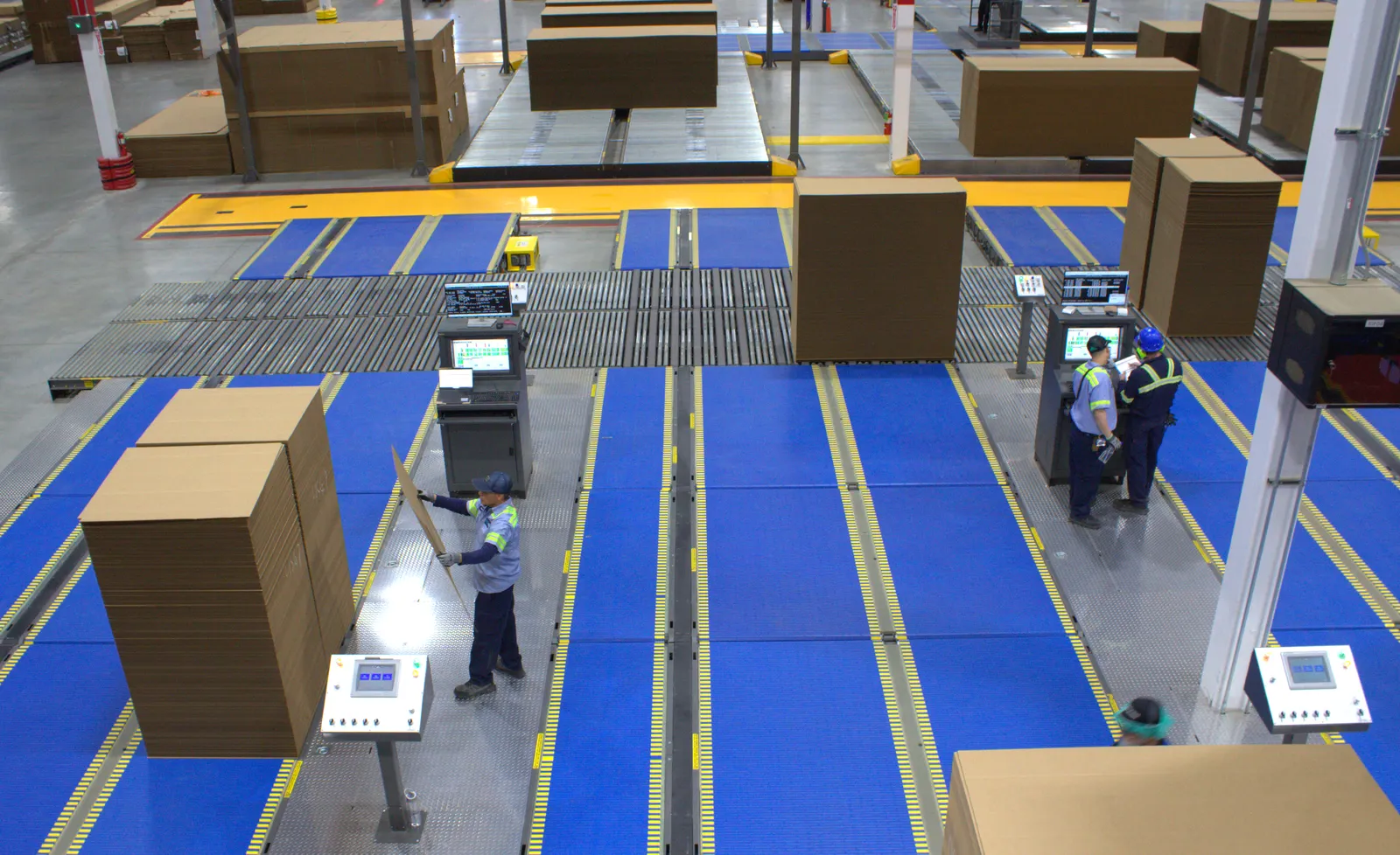 Employees examine stacks of corrugated boxes on conveyors in an industrial facility.