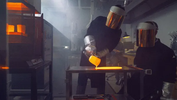 Two people wearing protective gear work with molten glass in an industrial lab setting.