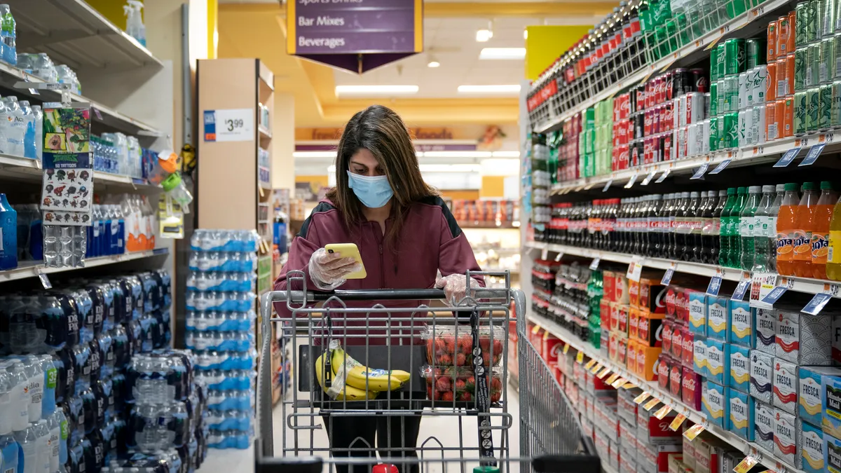 A person holding a smartphone pushes a shopping cart down a grocery aisle with bottled water on the left and soft drinks on the right.