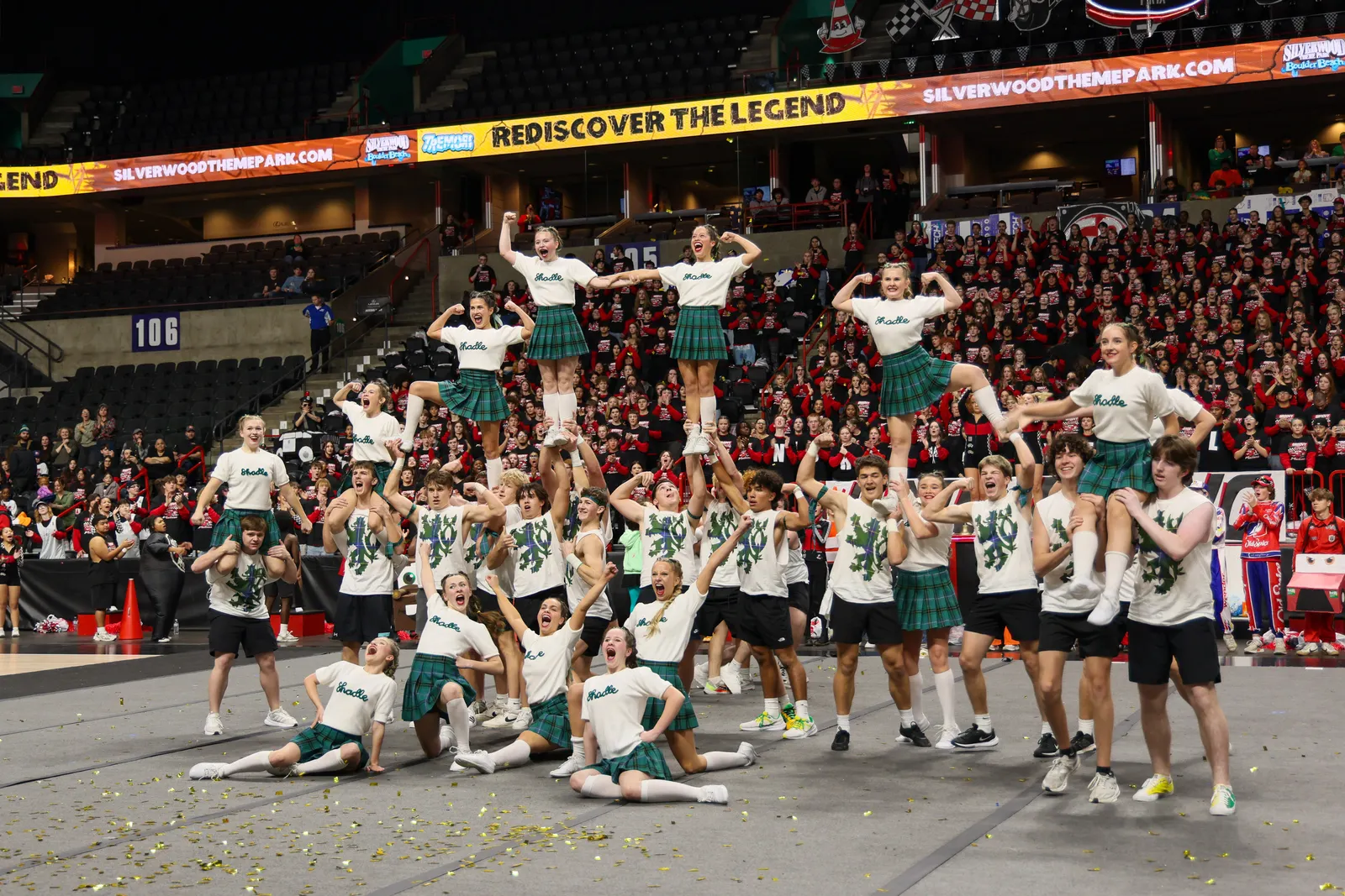 A group of people in uniforms form a human pyramid on a gym floor. Many spectators are watching from bleachers.