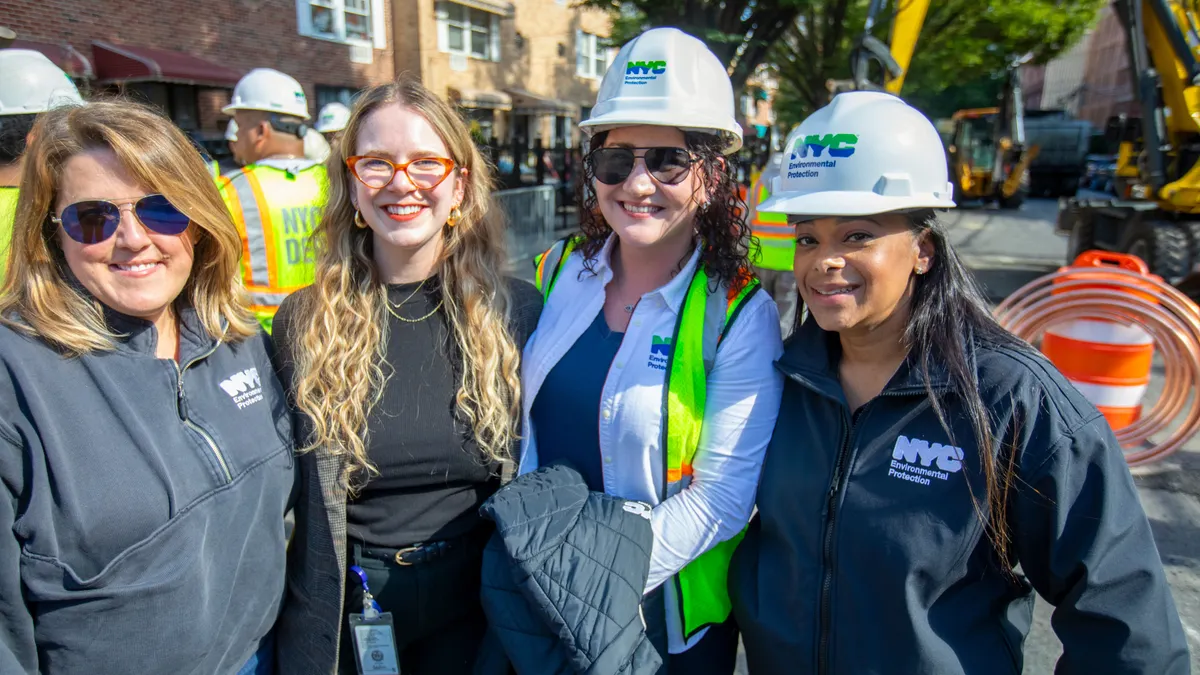 Four people standing together outdoors facing the camera, two with hard hats and three with NYC environmental protection logos on their tops. Behind them are workers in hard hats and safety vests.