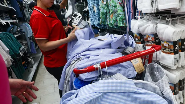 A woman in a red shirt uses a machine to put tags on a stack of shirts in a clothing section of a store.