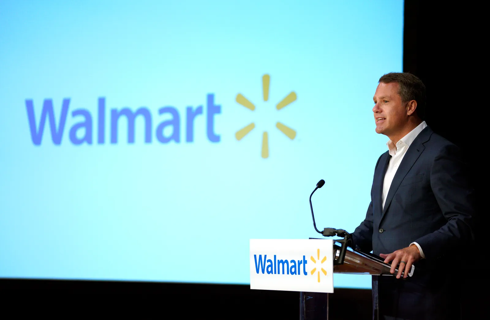 A business person speaking at a podium in front of a screen that says "Walmart"