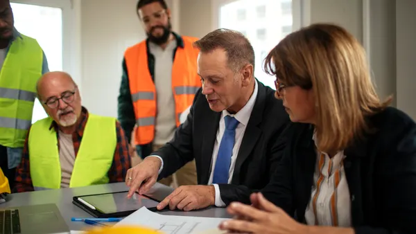A group of construction stakeholders, some in suits, some in work gear, gather in a meeting.