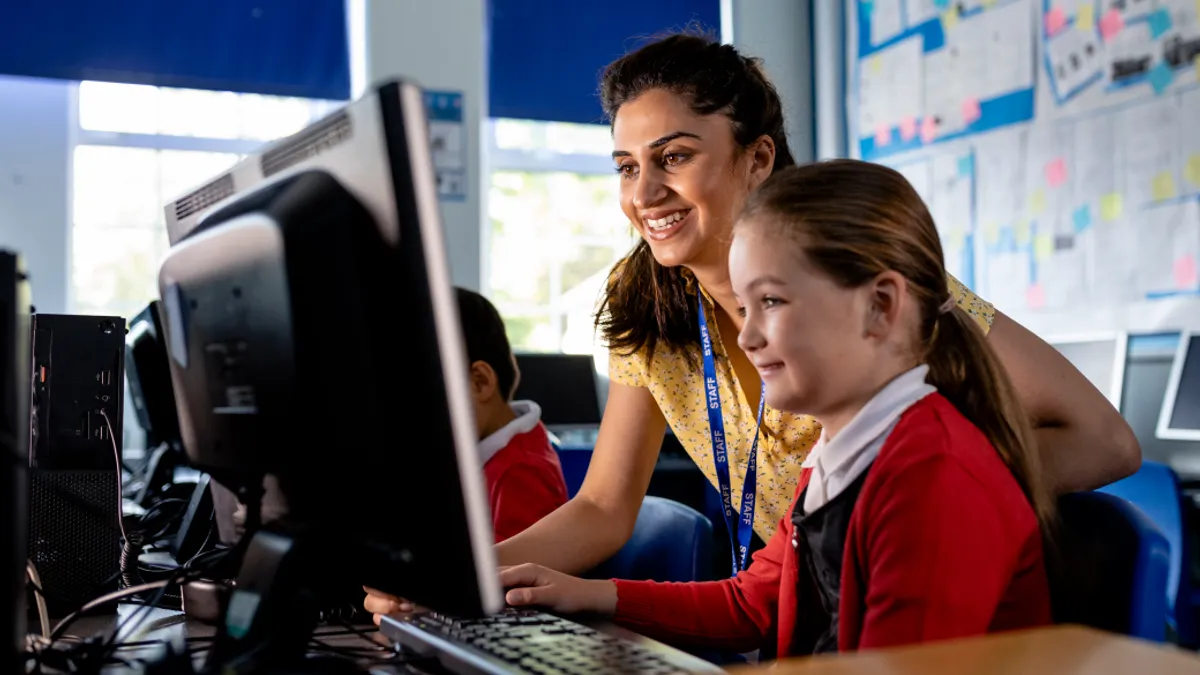 A grade school teacher assisting a student at a school desktop and smiling