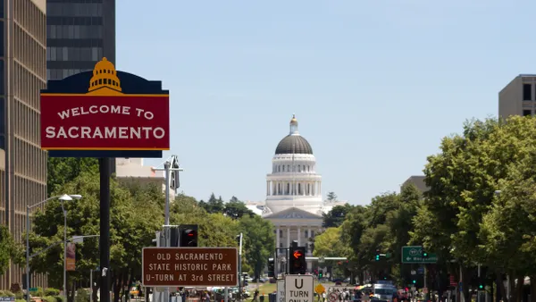 A sign that welcomes visitors to the city of Sacramento sits in the foreground, in front of a tall, white, domed building. Ahead is a long shot of grass, with buildings on the periphery.