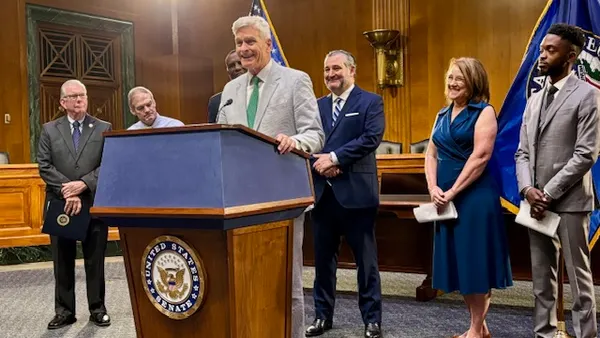 One person is standing behind a podium with a microphone in a room. Also pictured are five people standing behind the speaker and flags