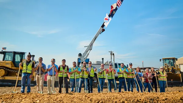 A group of people wearing high-visibility gear stand in front of shovels in the dirt on a bright day.