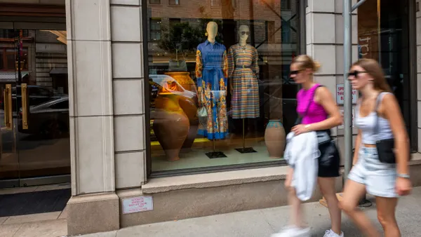 Two people walk in front of a store with a fashion display in the windows.