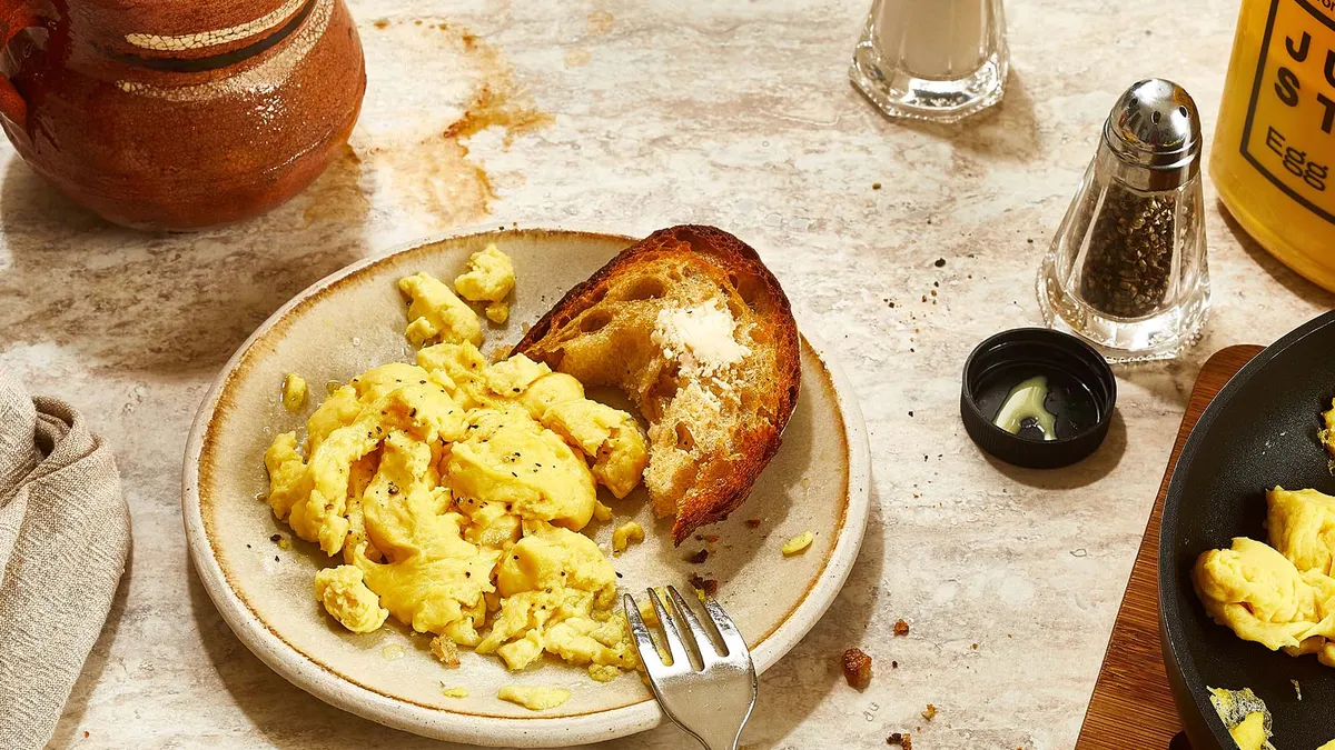 A small white plate of scrambled Just Egg and toast with a fork resting on it sits on a white marble surface. A coffee cup, salt and pepper shakers, a Just Egg bottle and skillet are nearby,
