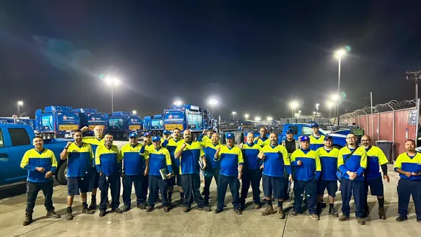 A group of waste collection workers stand in a truck yard, with trucks in background, under a dark sky