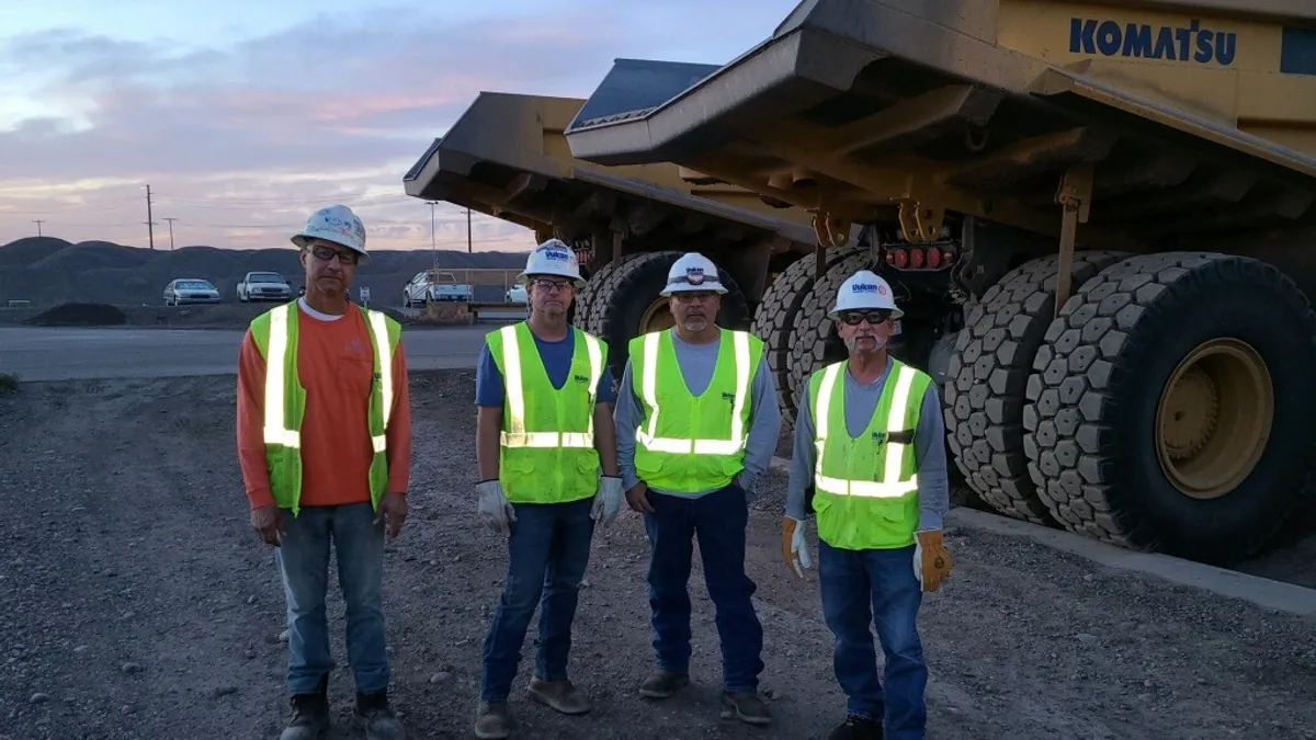 Four heavy machine operators wear hard hats and colorful vests as they stand in front of construction machinery.