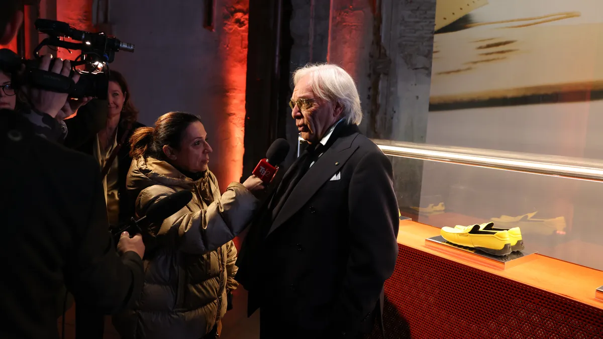 Diego Della Valle stands in front of a Tod's shoes display and speaks to a group of reporters hollding microphones and cameras.