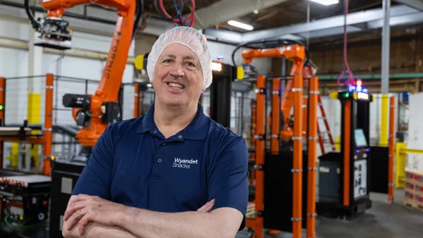 A man wearing a blue "Wyandot Snacks" polo shirt and a white hairnet stands smiling with arms crossed in a factory setting.