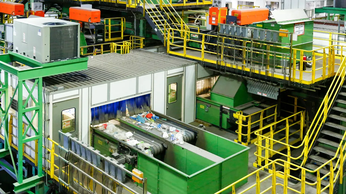 Plastic waste is carried out on conveyor belts in a modern industrial recycling plant. The interior is a maze of green and yellow metal structures, including stairs, bins and rooms.  