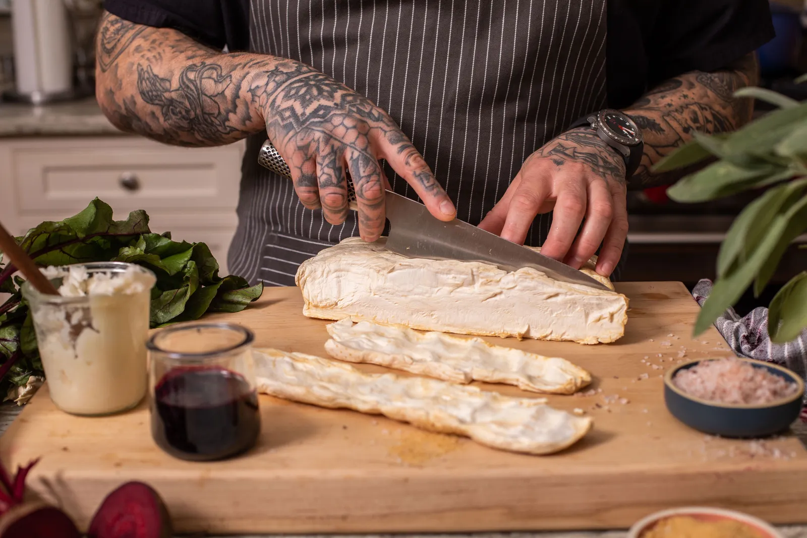 Someone wearing a striped apron uses a knife to slice a slab of mycelium on a wooden cutting board, with small containers of a white substance, something that looks like wine and salt on the board.
