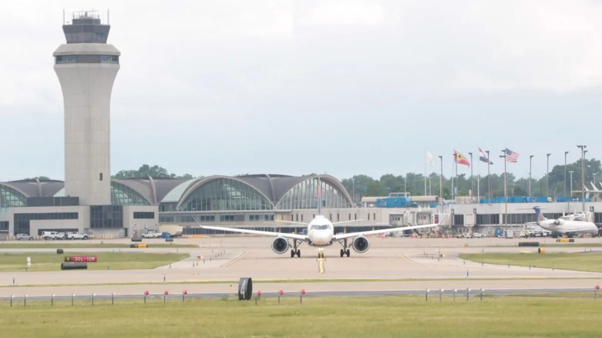 An airplane, facing the viewer, is positioned on a runway. In the background, other planes, flags and structures populate the scene.